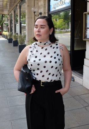 Blusa sin mangas blanca con lunares negros, cuello alto, combinada con pantalones negros de tiro alto y un bolso de cuero negro.