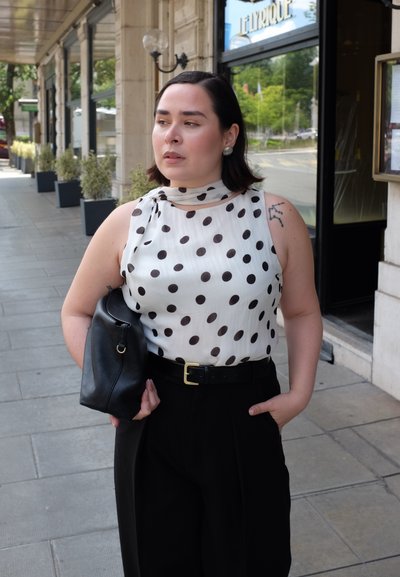Blusa sin mangas blanca con lunares negros, cuello alto, combinada con pantalones negros de tiro alto y un bolso de cuero negro.