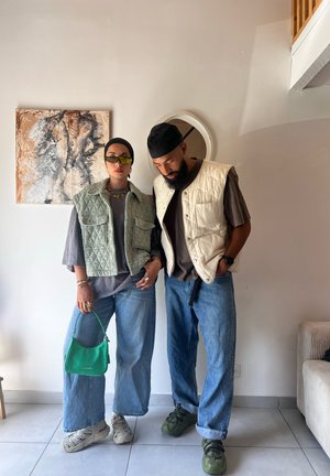 Two individuals stand side by side wearing layered outfits: a light green quilted vest, beige vest, gray top, loose jeans, and green handbag.