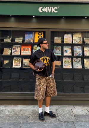 Camisa de malla negra con estampado dorado, pantalones cortos de patrón de leopardo, zapatos negros, calcetines blancos y un bolso marrón, de pie frente a una tienda de revistas.