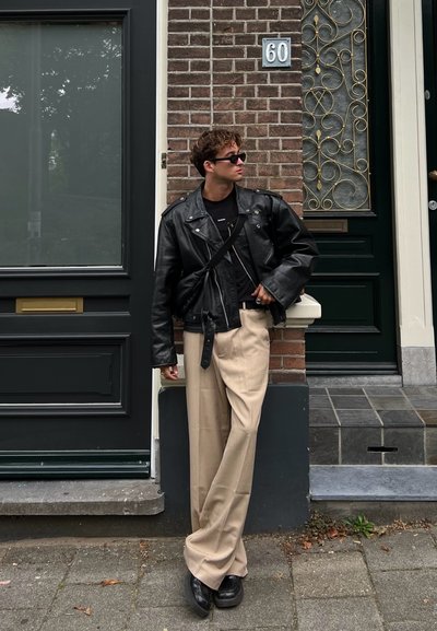 Chaqueta de cuero negra corta sobre una camiseta negra, pantalones de pierna ancha beige y zapatos de plataforma negros, con gafas de sol. Fondo de pared de ladrillo y puerta.