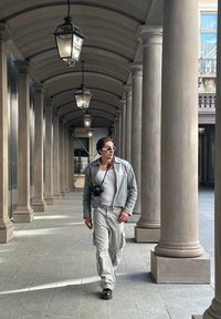 Γκρίζα δερμάτινη ζακέτα, λευκό tank top, ανοιχ�τό γκρίζοι cargo παντελόνια, μαύρες μπότες. Περπατώντας σε μια κιονοστοιχία με κρεμαστές φανάρια και πέτρινες κολώνες.