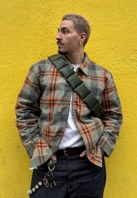 Chemise à carreaux vert, orange et bleu avec une fermeture éclair devant ; portée par-dessus un t-shirt blanc ; jean foncé ; sac épaule matelassé vert ; clés attachées.