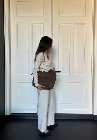 Bolso de cuero de ante marrón con una forma única, que presenta un asa superior y una textura suave, sostenido contra un fondo de puerta blanca. Llevado con pantalones de colores claros.