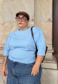 Pulover tricotat de culoare albastru deschis, cu mâneci scurte puff, asociat cu o fustă din denim. Include o geantă din piele maro și o șapcă maro.
