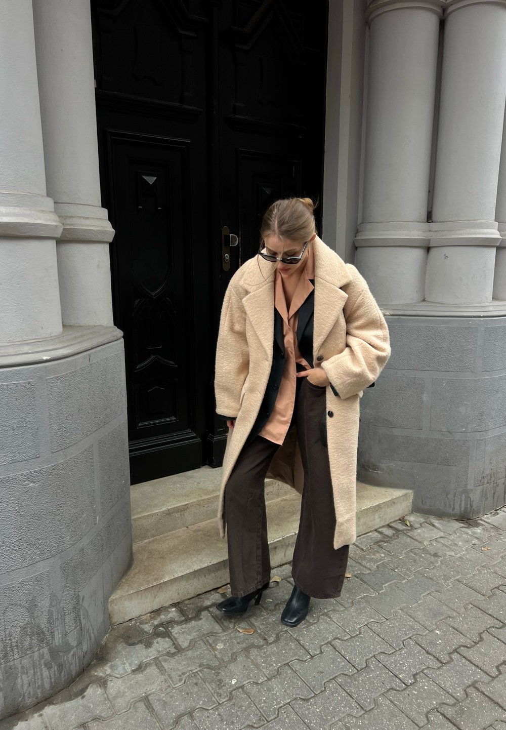 Femme portant un long manteau beige, une chemise pêche, un blazer noir et un pantalon sombre, debout sur des marches à l'extérieur d'un bâtiment avec de grandes colonnes grises et des portes noires.
