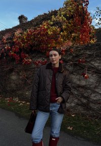 Hnedá prešívaná bunda cez bordový sveter, modré džínsy a červené gumáky; drží tmavú kabelku na pozadí farebného jesenného lístia.