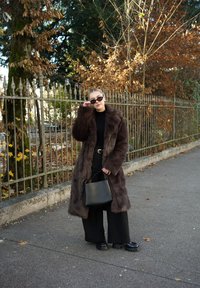 Cappotto in pelliccia sintetica marrone, dolcevita nero, pantaloni neri a gamba larga, occhiali da sole rotondi neri e borsa a secchiello nera in una strada con foglie autunnali.