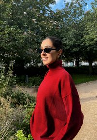 Maglione rosso a collo alto lavorato a maglia, con una vestibilità comoda, caratterizzato da un colletto a coste. Sono presenti anche occhiali da sole neri e un ambiente naturale all'aperto.