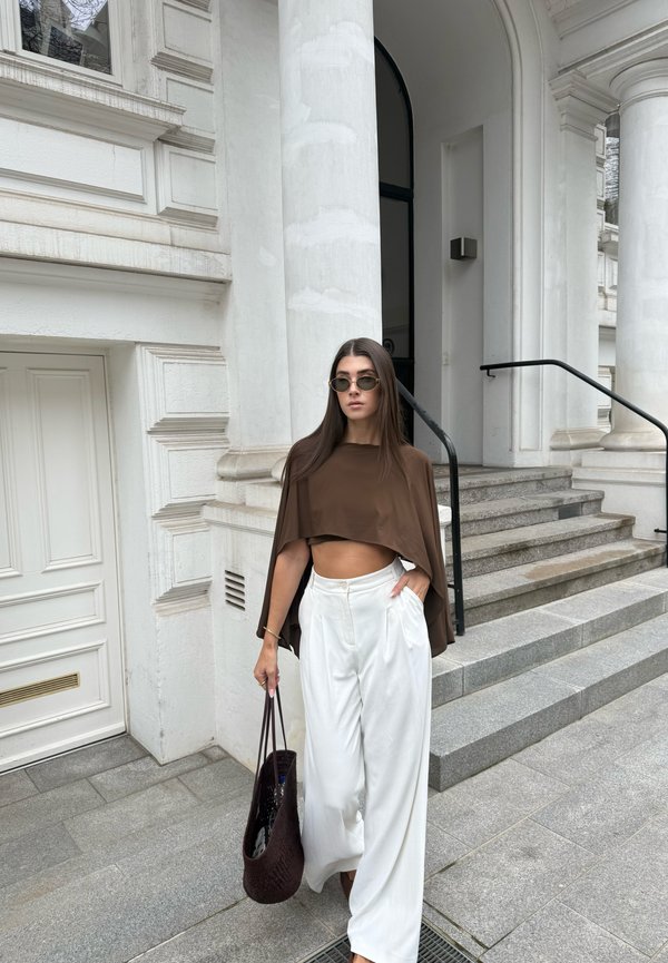 Žena nosiaca slnečné okuliare, hnedý krátky top a široké biele nohavice kráča pri schodoch bielej budovy s stĺpmi a drží tmavú pletenú tašku.