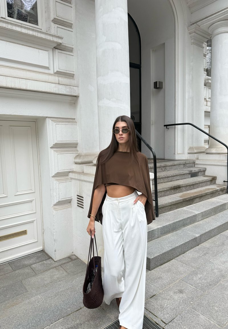 Γυναίκα με γυαλιά ηλίου, καφέ κοντό τοπ και φαρδιά λευκά παντελόνια περπατά κοντά σε σκαλοπάτια κτηρίου με λευκούς κίονες, κρατώντας σκούρα πλεκτή τσάντα.