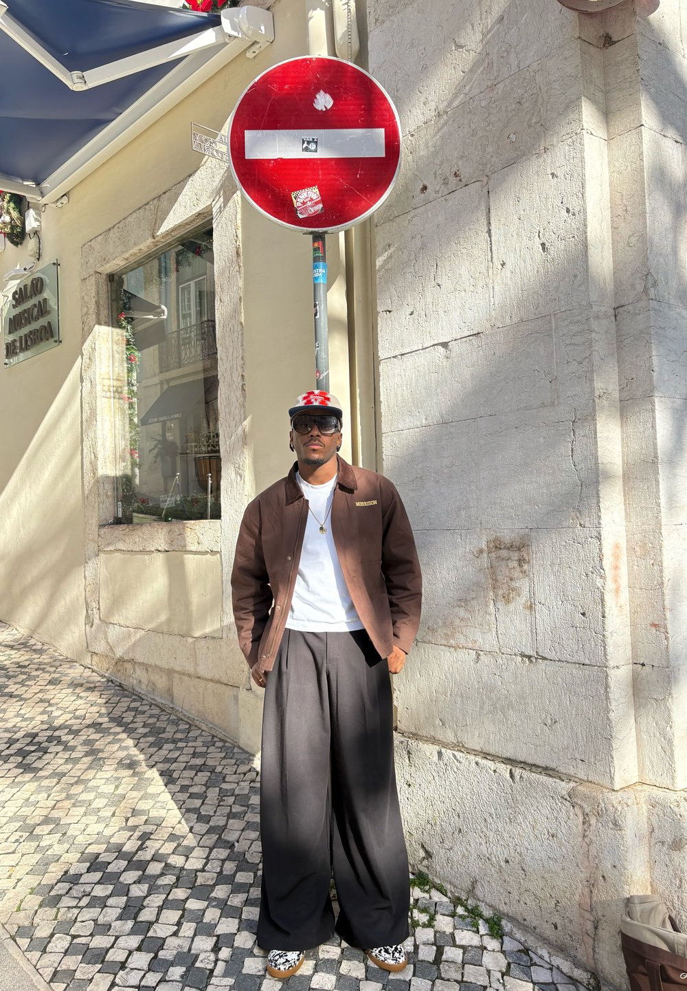 Pantalon noir à jambes larges, t-shirt blanc, veste marron. Debout sous un panneau de sens interdit fixé sur un mur en pierre dans une rue ensoleillée.
