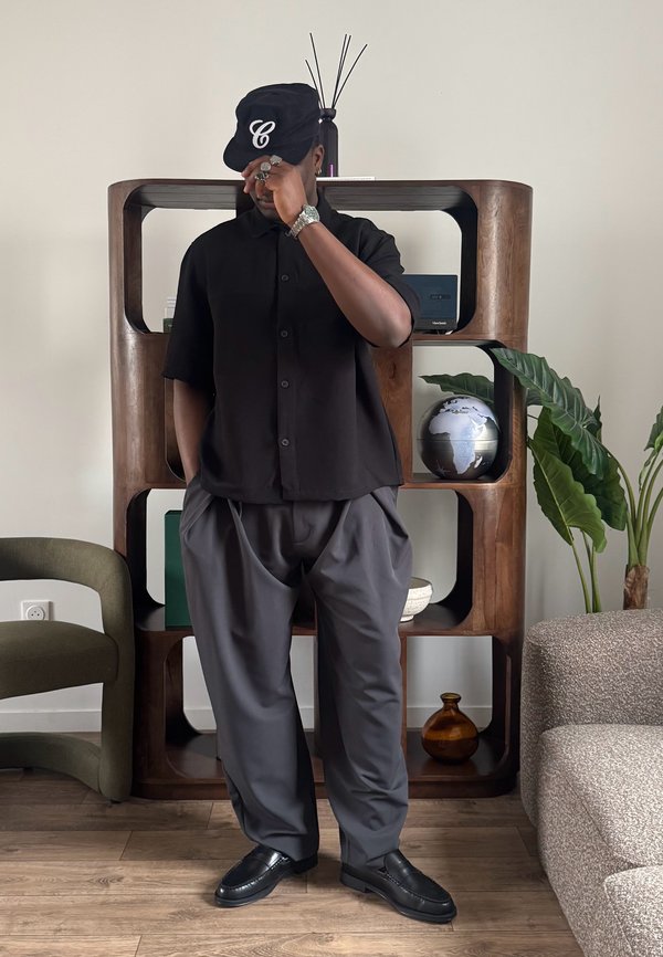 Bărbat cu șapcă neagră și cămașă cu pantaloni gri stă în fața unei rafturi de lemn ținând viziera șepcii în interior.