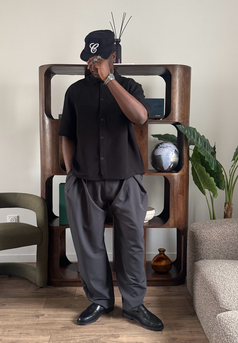 Homme portant une casquette noire et une chemise avec un pantalon gris, se tenant devant une étagère en bois et tenant la visière de sa casquette à l'intérieur.