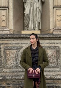 Femme en manteau vert olive tenant un sac à main bordeaux, debout devant un mur de pierre orné avec une statue partiellement visible au-dessus.