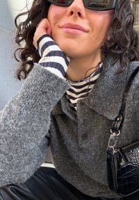 Γυναίκα με σγουρά σκούρα μαλλιά που φοράει μαύρα γυαλιά ηλίου, γκρι υφασμάτινο παλτό πάνω από μαύρη και λευκή ριγέ μπλούζα, στηρίζει το κεφάλι της στο χέρι, κρατώντας μαύρη τσάντα.