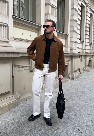 Hombre con chaqueta marrón, camisa negra, pantalones blancos, zapatos negros y gafas de sol, sosteniendo un bolso negro, de pie en la acera de la ciudad junto a un edificio ornamentado.