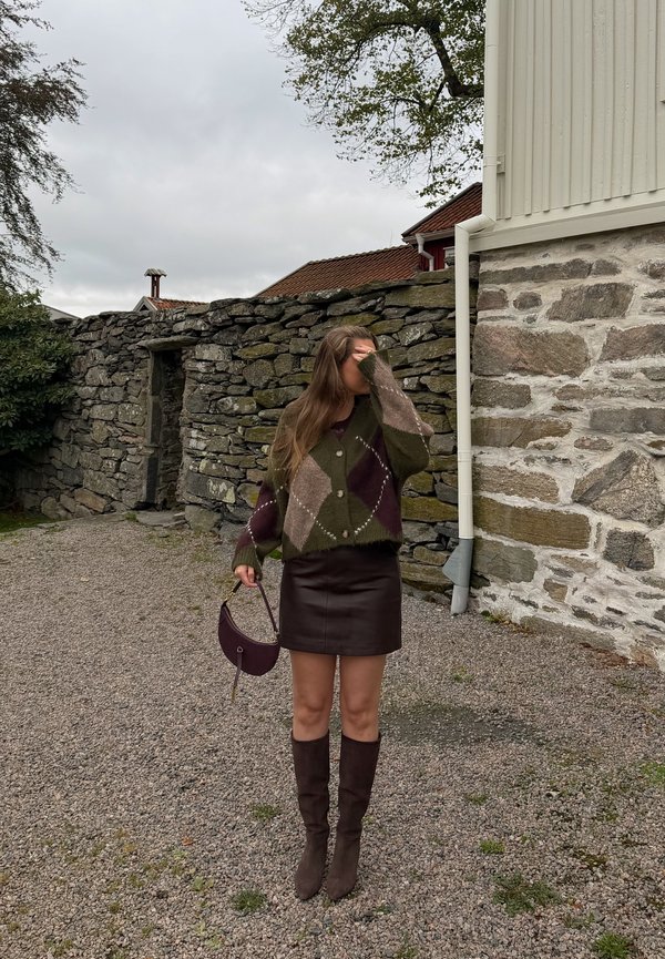 Una mujer con un jersey estampado, una falda oscura y botas hasta la rodilla sostiene un bolso junto a un muro de piedra y se cubre la cara con una mano.