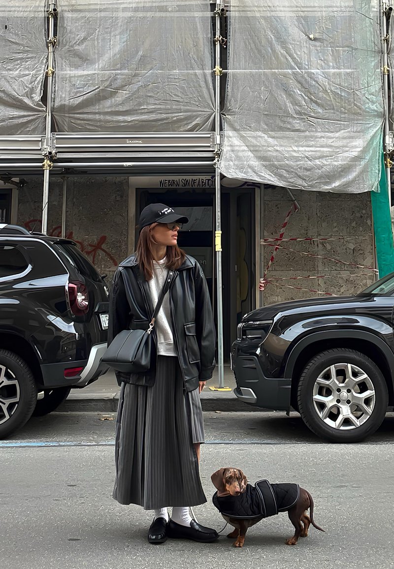 Casaco de couro preto sobre sweatshirt branca, saia cinzenta plissada, sapatos pretos e mala. Um pequeno cão castanho com um casaco preto está próximo.