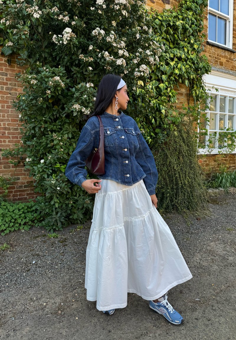 Femme en veste en jean, jupe blanche à volants, baskets bleues et boucles d'oreilles en forme de cœur, debout à l'extérieur près de vignes vertes fleuries et d'un bâtiment en briques.