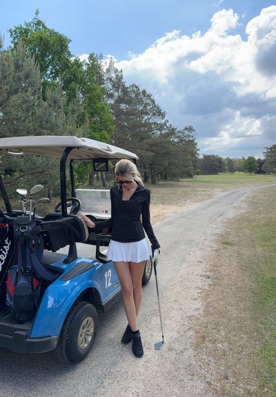 Carrito de golf azul con acentos beige aparcado en un camino. Persona con camiseta negra y falda blanca sosteniendo un palo de golf, con árboles de fondo.