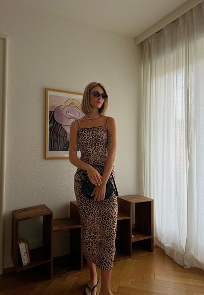 Vestido midi de estampado de leopardo con tirantes finos, fondo rosa claro y manchas oscuras; complementado con bolso negro y gafas de sol.