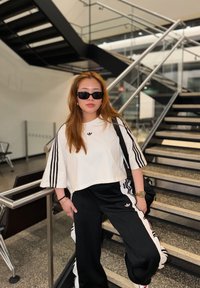 Jeune femme aux longs cheveux portant des lunettes de soleil noires, un t-shirt blanc Adidas, un pantalon de survêtement noir Adidas et une montre en or, debout près d'un escalier intérieur.