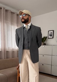 Homme portant une casquette beige, des lunettes, un blazer gris, un pull gris, une chemise blanche, une cravate rayée et un pantalon beige, debout dans un salon avec des rideaux beiges.