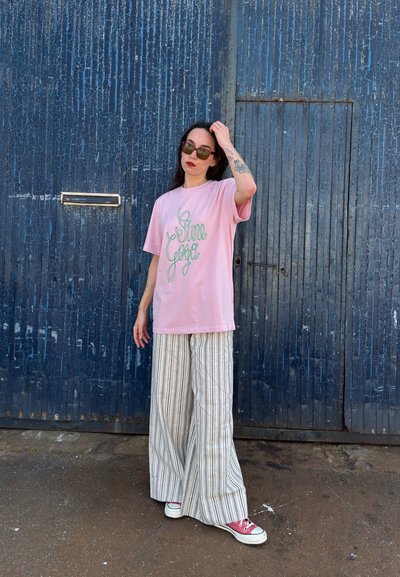 Camiseta de algodón rosa con texto en cursiva verde, combinada con pantalones ligeros de rayas anchas. Gafas de sol negras y zapatillas rojas completan el look.