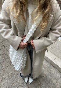 Beżowy wełniany płaszcz noszony na białym swetrze dzianinowym, trzymając fakturowaną białą torebkę. Stylizacja obejmuje szare dżinsy i białe szpilki w szpic.