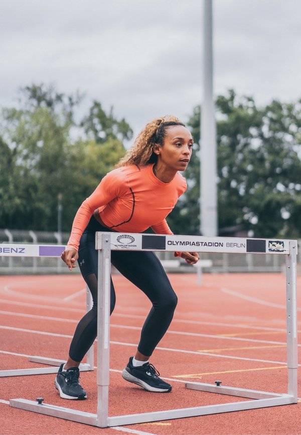 Athlète en haut orange et leggings noirs se préparant à sauter une haie sur une piste extérieure à l'Olympiastadion de Berlin.