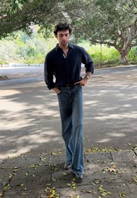 Granatowa koszula z długim rękawem, z podwiniętymi rękawami, luźne jasnoniebieskie dżinsy, czarny pasek i ciemne buty, idący po ścieżce usłanej liśćmi.