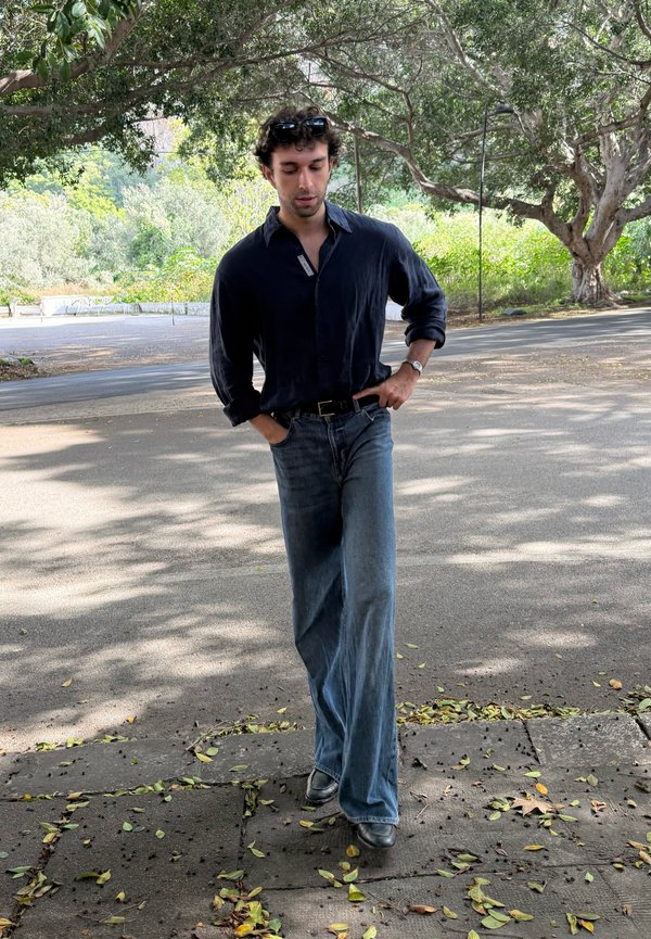 Dark blue button-up shirt with rolled sleeves, loose light blue jeans, black belt, and dark shoes, walking along a path covered in leaves.
