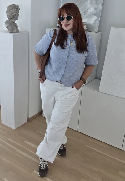 Suéter de punto gris claro con botones, pantalones anchos blancos, zapatillas marrones y gafas de sol. El fondo presenta una decoración minimalista y una escultura.