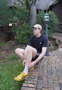 Jeune homme en tenue de sport noire et casquette beige assis sur un muret en briques en plein air, ajustant sa chaussette blanche et portant des baskets jaunes.