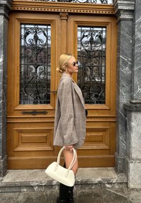 Gestreepte oversized beige blazer, zwarte kniehoge laarzen en een kleine witte handtas. Getextureerde houten deur met uitgebreide ijzeren bewerking op de achtergrond.