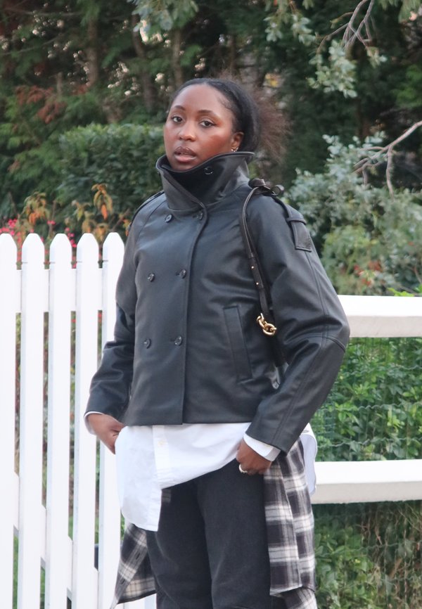 Czarna, dwurzędowa kurtka ze skóry z wysokim kołnierzem, zestawiona z białą koszulą i ciemnymi dżinsami. Kurtka ma pagony na ramionach i jest uzupełniona torebką.