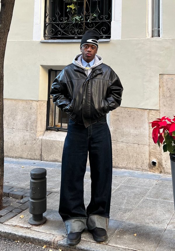 Čierna kožená bomber bunda cez šedú mikinu, skombinovaná s nadmernými modrými džínsami a čiernymi topánkami. Stojí na mestskom námestí.