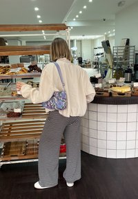 Χαλαρή λευκή μπλούζα με μακριά μανίκια, μαύρο και άσπρο ριγέ παντελόνι με φαρδιές γραμμές και μια μπλε τσάντα με λουλούδια. Προθήκη με ψημένα προϊόντα στο background.