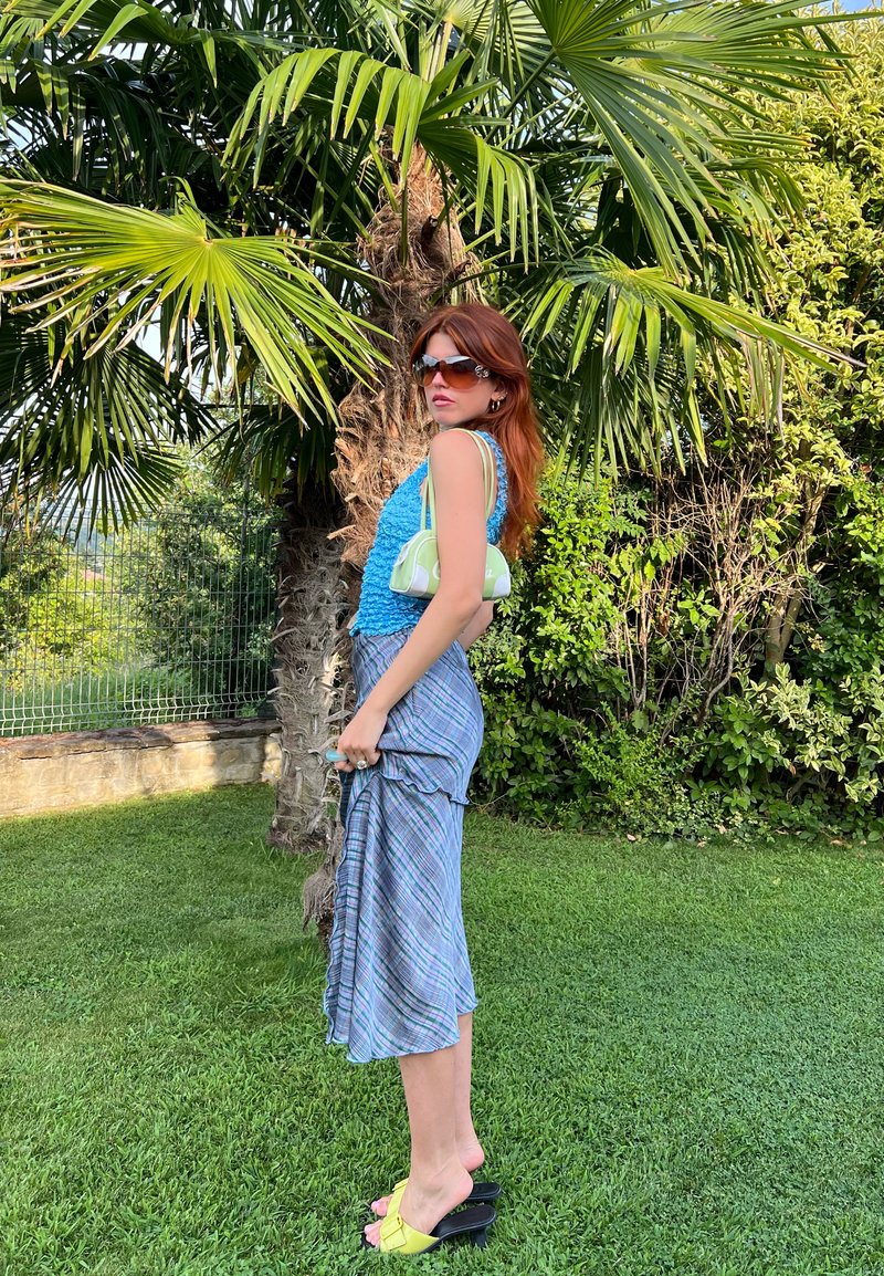 Ανοιχτό μπλε υφασμάτινη μπλούζα, καρό μίντι φούστα σε αποχρώσεις μπλε και πράσινου, κίτρινα ψηλά τακούνια και μικρή πράσινη τσάντα, σε φόντο πλούσιας φυτολογίας.