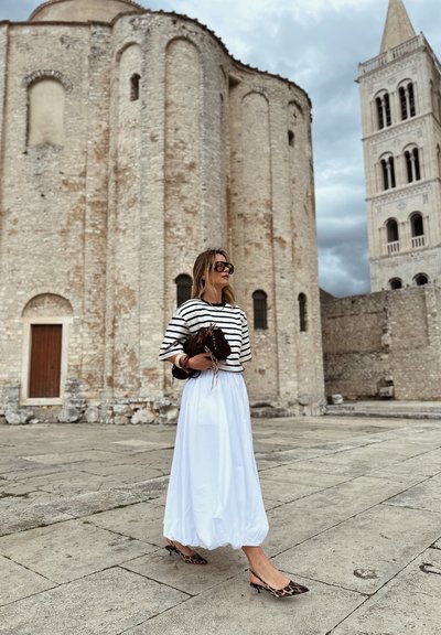 Blusa a rayas blancas y negras, falda maxi blanca y zapatos slingback de leopardo. De pie junto a un edificio de piedra con una puerta de madera.
