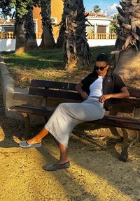 Γυναίκα καθισμένη σε ένα ξύλινο παγκάκι, φορώντας μαύρη κοντομάνικη μπλούζα, λευκό φανελάκι, γκρίζα φούστα και μαύρα παπούτσια με λευκές βούλες.