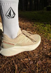 Adidași Salomon bej cu un suprafețe texturate, talpă albă cu accente decorative, și șireturi ușoare, purtați cu ciorapi albi care au un logo negru.