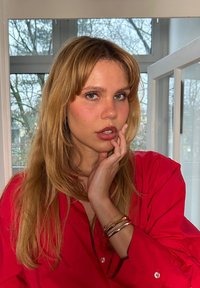 Une personne portant une chemise rouge à boutons se pose avec une main sur le visage, mettant en avant de longs cheveux blonds et des bracelets en argent à son poignet.