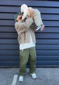 Ένας άντρας σε καφέ χνουδωτή ζακέτα κρατά ένα παιδί που φοράει μπεζ φούτερ και πράσινα cargo παντελόνια. Είναι μπροστά σε σκούρο μπλε φόντο.