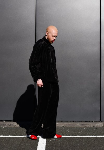 Chándal de terciopelo negro con chaqueta de cremallera y pantalones holgados, combinado con calzado rojo y blanco, sobre un fondo gris.