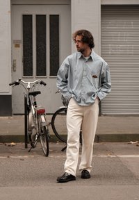 Ριγέ πουκάμισο σε ανοιχτό μπλε και λευκό, συνδυασμένο με κρέμ παντελόνι και μαύρα παπούτσια, στέκεται κοντά σε ένα ποδήλατο σε έναν δρόμο.