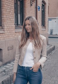 Beige jakke med gulldetaljer over en hvit topp, kombinert med blå høyhalsede jeans. Langt, rett hår rammer ansiktet.