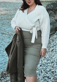 Maglione avvolgente bianco a maglia con costine spesse e una cintura in vita, abbinato a una gonna aderente verde oliva. Tieni un giubbotto oliva, in piedi su ciottoli.