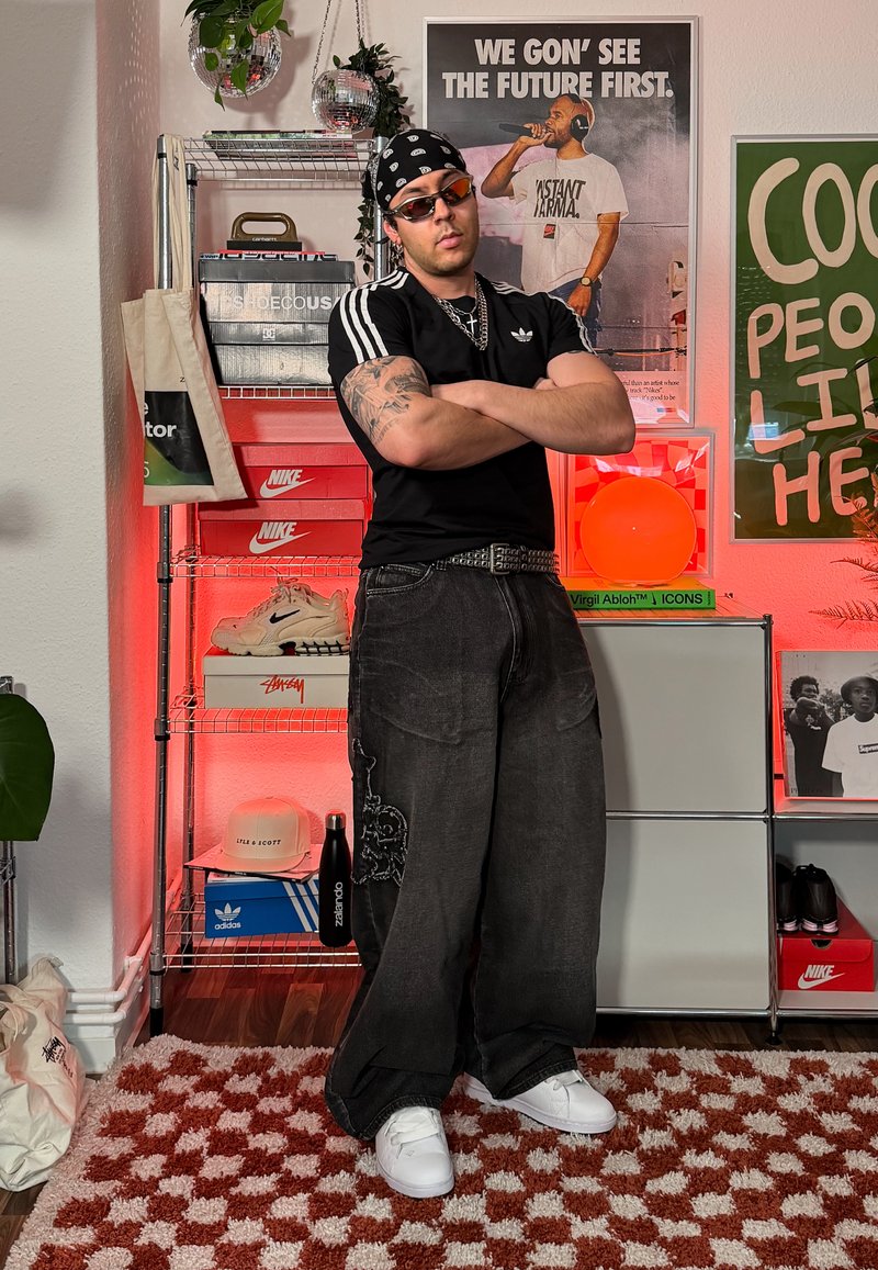 Homme avec des tatouages portant un t-shirt Adidas noir et un bandana, debout les bras croisés dans une pièce avec des boîtes de baskets et des affiches sur le mur.